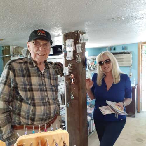 A man and a woman smile in a shop, surrounded by crafts and accessories, with a wooden post in the center.