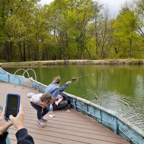 A child fishing from a dock while another child watches, surrounded by trees and a calm green lake.