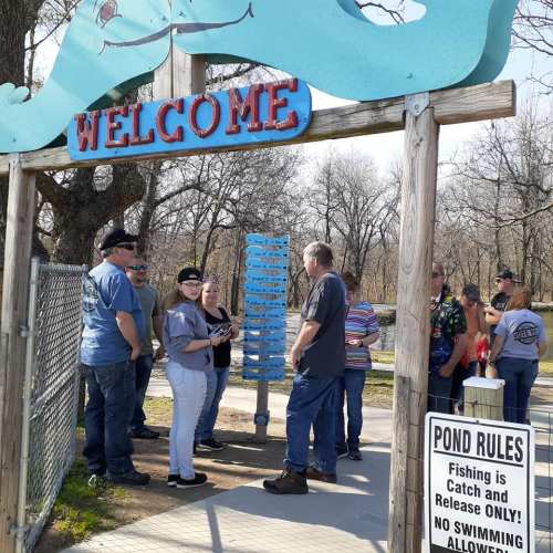A group of people gathered at a welcome sign near a pond, discussing rules and enjoying a sunny day outdoors.