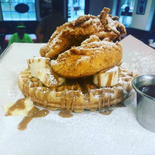 A plate featuring a waffle topped with fried chicken, banana slices, powdered sugar, and a drizzle of syrup.