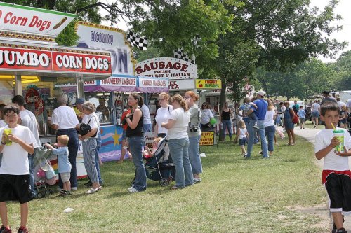 A busy fair scene with food stalls, families, and children enjoying the festivities on a sunny day.