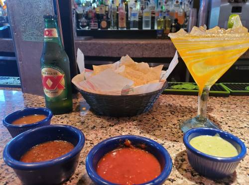 A table with a basket of tortilla chips, four bowls of salsa, a beer, and a margarita in a bar setting.