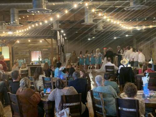 A wedding ceremony in a rustic venue with guests seated, a bridal party in blue dresses, and string lights overhead.