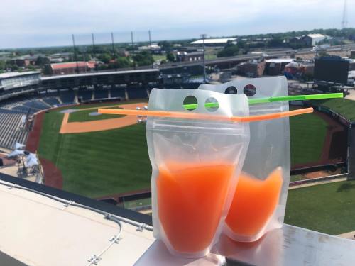 Two colorful drinks in pouches with straws, overlooking a baseball field and stadium in the background.