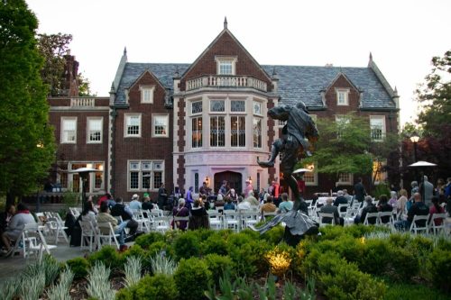 A garden event with guests seated in white chairs in front of a large brick mansion and a statue, surrounded by greenery.