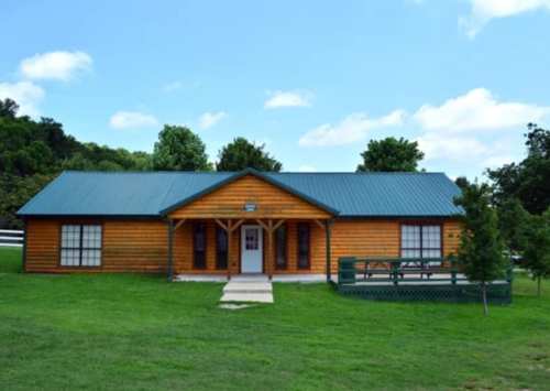 A wooden cabin with a blue metal roof, surrounded by green grass and trees under a clear blue sky.