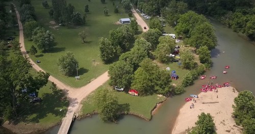 Aerial view of a green park with a river, camping area, and people kayaking near a sandy beach.