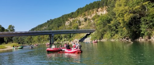 A group of people in red inflatable boats paddling on a calm river under a bridge, surrounded by green hills.