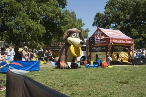 A large inflatable dog mascot stands in a park during a dog event, with spectators and vendor tents in the background.
