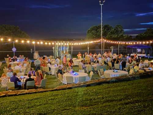 A lively outdoor event at night with guests dining at tables under string lights, surrounded by greenery.
