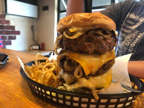 A towering burger with two patties, melted cheese, grilled onions, and lettuce, served with curly fries in a basket.