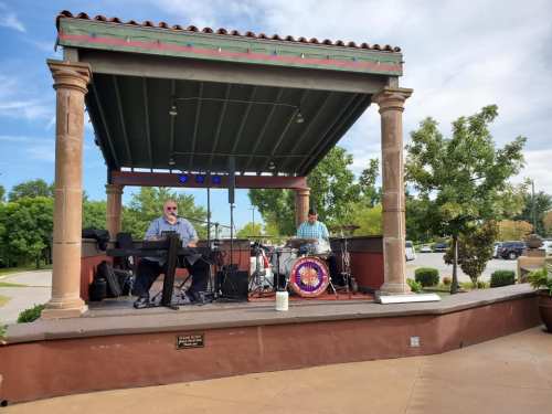 Two musicians perform on a stage with columns, featuring a keyboard and drums, set in a park on a sunny day.