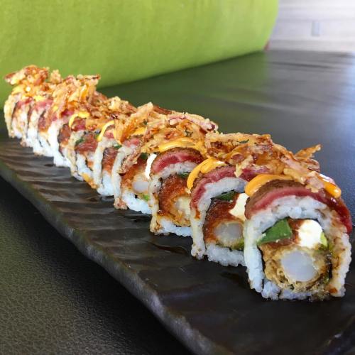 A close-up of a sushi roll topped with crispy onions and drizzled with sauce, served on a black plate.