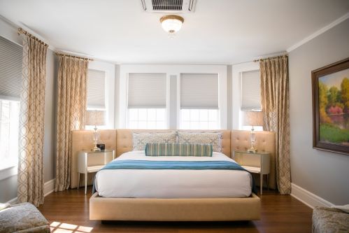Bright, modern bedroom with a large bed, decorative pillows, and elegant curtains by windows, featuring soft lighting.