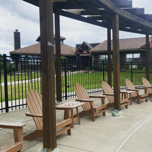 A row of wooden Adirondack chairs under a pergola, with a grassy area and a building in the background.