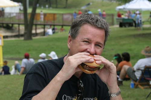 A man smiles as he prepares to take a big bite of a hamburger at an outdoor event.