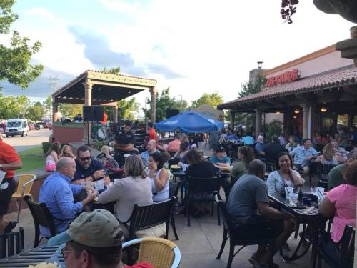 A lively outdoor dining scene with people enjoying food and drinks at tables under a blue sky.