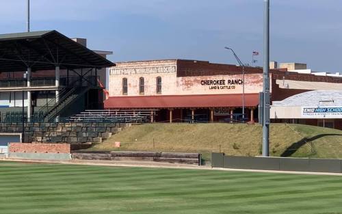 View of a baseball field with bleachers and a building labeled "Cherokee Ranch Land & Cattle Co" in the background.