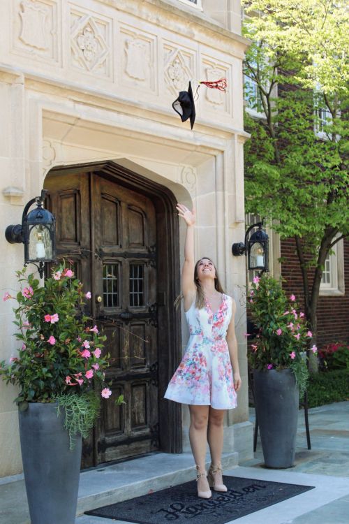 A graduate in a floral dress throws her cap in the air in front of a beautiful building with flowers.