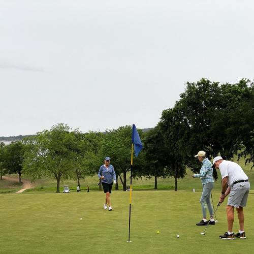 Three golfers on a green, one putting near the hole, with trees and a cloudy sky in the background.