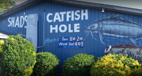 A blue building with a mural of a catfish and a sign displaying hours for Shad's Catfish Hole.