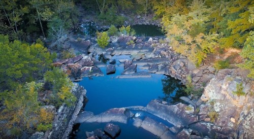Aerial view of a serene natural pool surrounded by rocks and trees, reflecting the clear blue sky.