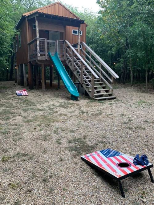 A treehouse with a slide, surrounded by trees, and two cornhole boards featuring American flags on the ground.