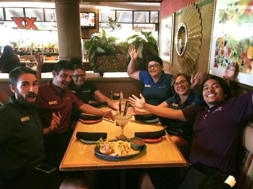 A group of six restaurant staff members smiling and posing with hands raised at a table with food and drinks.