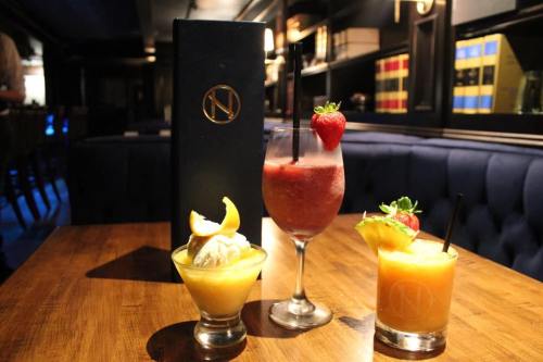 Three colorful cocktails on a wooden table, with a menu in the background, set in a stylish bar.