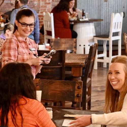 A smiling waitress takes an order from two happy customers at a cozy restaurant with wooden tables.