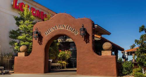 A welcoming archway with "Bienvenidos" sign, leading to a restaurant surrounded by greenery and blue sky.