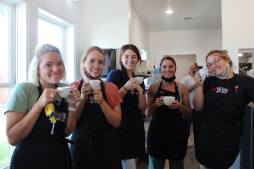 A group of five smiling individuals in aprons hold cups, standing in a bright café setting.