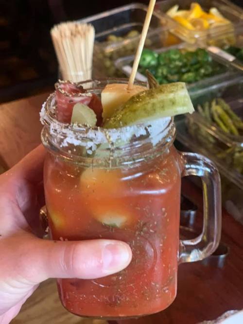 A hand holding a mason jar filled with a colorful cocktail garnished with pickles, meats, and a salt rim.