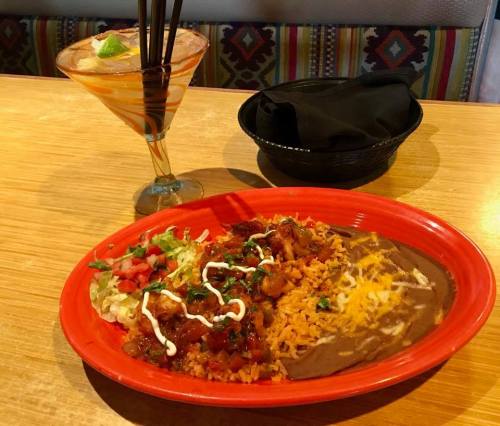 A vibrant plate of rice, beans, and salad topped with sauce, alongside a colorful cocktail on a wooden table.