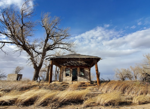 Abandoned building with a wooden porch, surrounded by dry grass and bare trees under a cloudy blue sky.
