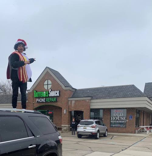 A person in a festive outfit stands on top of a car outside a phone repair shop, with snow falling around them.