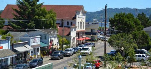 A quaint street lined with shops and trees, featuring parked cars and a view of distant hills.
