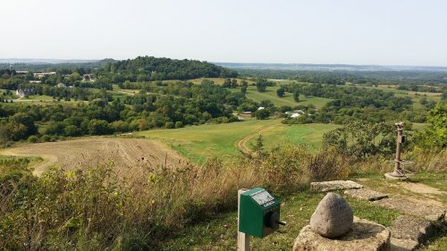 A scenic view of rolling green hills and fields under a clear sky, with a stone marker in the foreground.