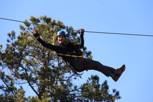 A person in a blue helmet zip-lining through trees against a clear blue sky, smiling and enjoying the adventure.