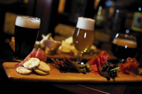 A wooden platter with assorted meats, cheeses, and crackers, accompanied by three glasses of different beers.