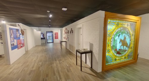 A spacious gallery with white walls, wooden flooring, and a large stained glass display at the end of the hallway.