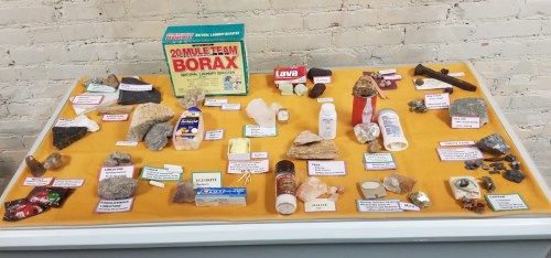 Display of various minerals and rocks, including Borax, lava, and labeled samples on a yellow tablecloth.