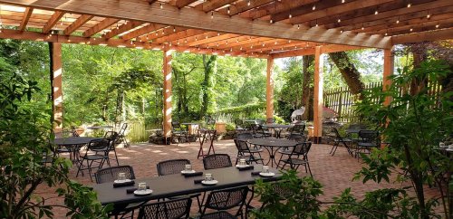 Outdoor dining area with black tables and chairs, surrounded by greenery and wooden beams, illuminated by string lights.