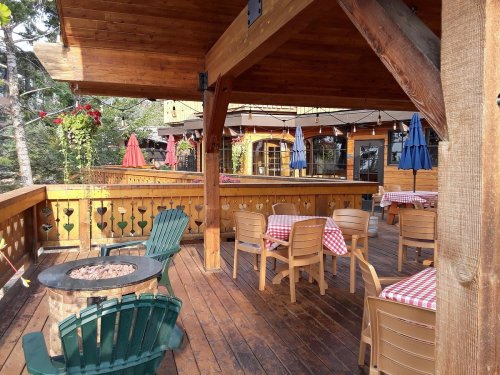 A cozy wooden deck with checkered tablecloths, chairs, and umbrellas, surrounded by greenery and rustic decor.