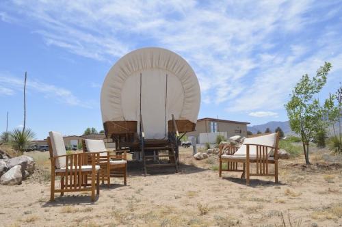A covered wagon sits on a sandy area, surrounded by wooden chairs and sparse vegetation under a blue sky.