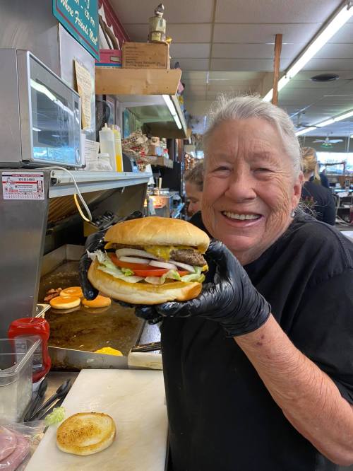 A smiling woman in a black shirt holds a large burger with lettuce, tomato, and onion in a diner kitchen.