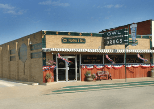 A brick building with a sign for "Owl Prescriptions Drugs" and "Soda Fountain & Grill," decorated with red, white, and blue bunting.