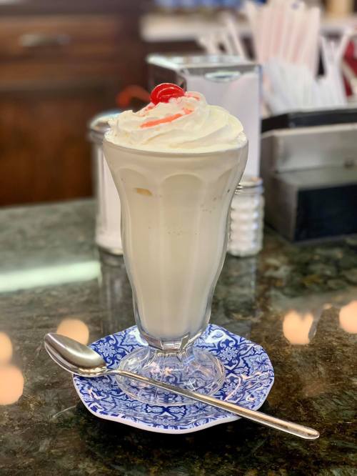 A tall glass of vanilla milkshake topped with whipped cream and a cherry, served on a decorative blue plate.