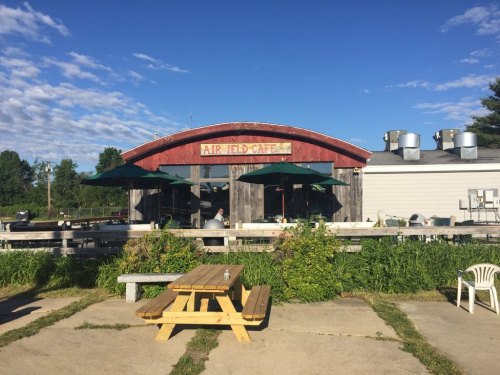 A rustic café with a red arched roof, outdoor seating, and green umbrellas, surrounded by greenery under a blue sky.