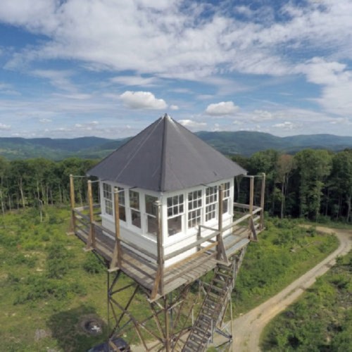 A tall observation tower with a peaked roof, surrounded by trees and mountains under a blue sky with clouds.